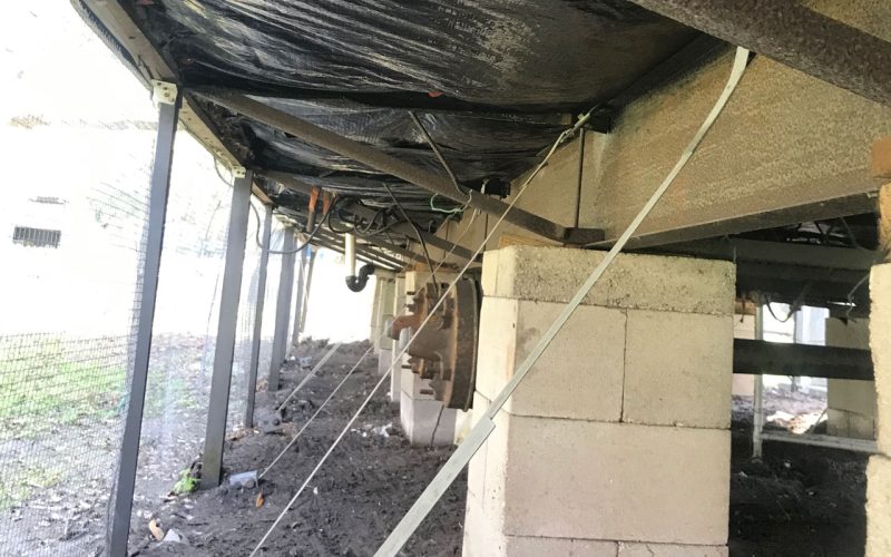 View inside a crawl space showing black vapor barrier installed on ceiling joists, concrete piers, and steel tie-down straps running diagonally — illustrating mid-project progress before full encapsulation.