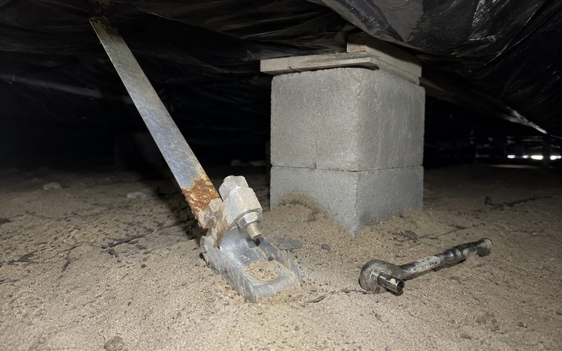 A corroded tie-down anchor head at ground level beneath a manufactured home, showing rust before replacement.