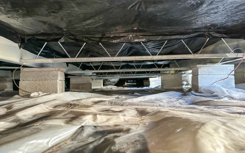 Tightly installed black vapor barrier under floor joists, anchored with orange fasteners, creating a seamless moisture barrier across the entire crawl space ceiling for improved home air quality and structural integrity.