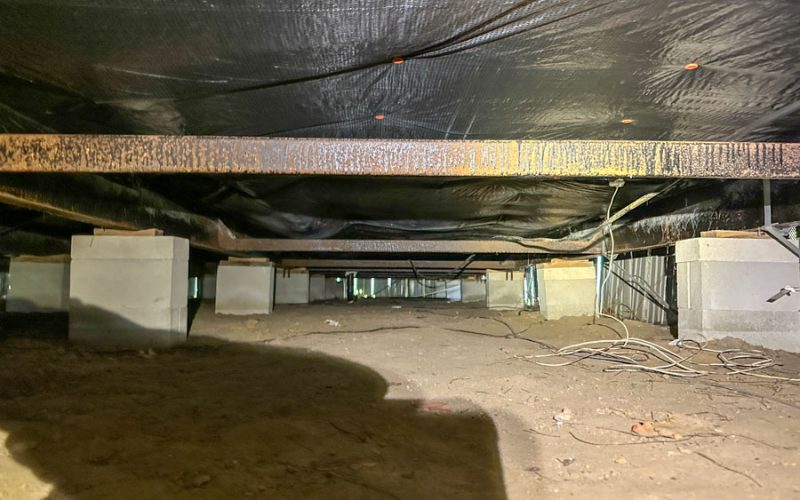 Wide-angle shot of an unencapsulated crawl space featuring exposed steel I-beams, concrete piers, and a dirt floor — illustrating the pre-installation condition before East Coast Vapor Barrier’s moisture control system is applied.